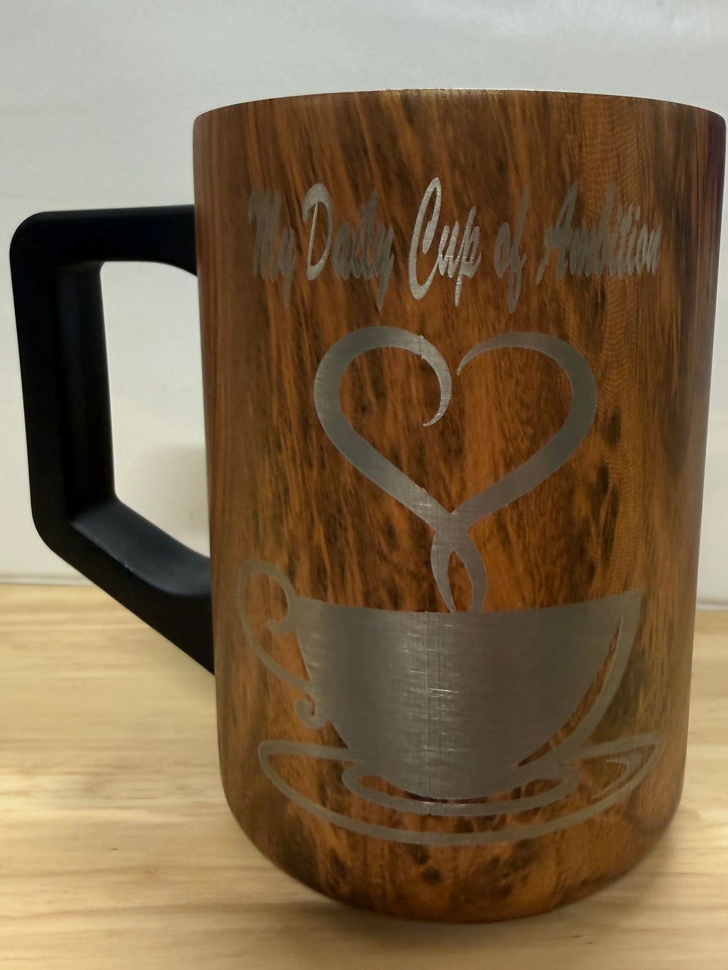 A wooden mug with a black handle, featuring a laser-engraved design of a coffee cup and the text 'The Daily Cup of Ambition' 

